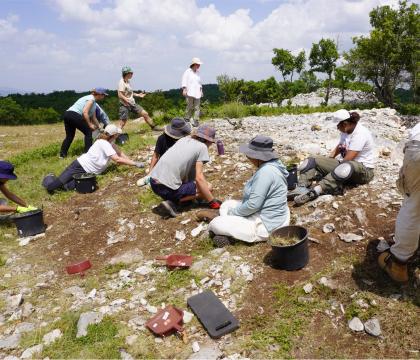 Several archaeologists sit on the ground excavating a site on a stony and grassy hillside against a cloudy blue sky.