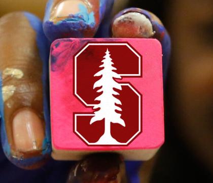 Paint covered hand holding a square block with the Stanford S insignia