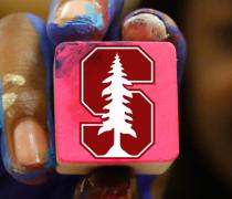 Artist's fingers covered in paint, presenting a stamp block with the Stanford tree logo.