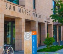 Exterior view of the San Mateo Public Library building on a sunny day, showing the stone facade with the library name, a sidewalk lined with trees, street banners, bicycle racks, and nearby parking signage.