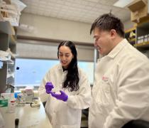 Mentor and mentee in lab coats working together at a laboratory bench. DRIVE student is holding a small dish and using a tool while the mentor stands beside them observing. The workspace includes lab supplies, equipment, and shelves in the background.