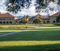Sunlit university quad with green lawn, arched sandstone buildings, and red tile roofs.